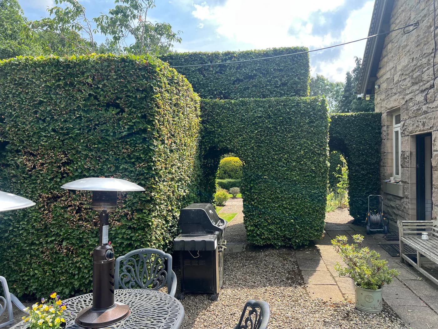 A neatly trimmed tall hedge with an archway cut through it, beside a garden patio